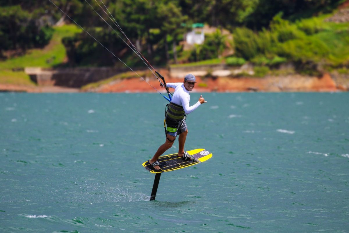 Kitefoiler riding across flat green water below a wooded shore